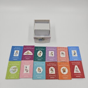 May include: A collection of twelve small Beatrix Potter books arranged in two rows, including titles like The Tale of Peter Rabbit and The Tale of Tom Kitten, accompanied by an open, empty white cardboard storage box with a decorative illustration.