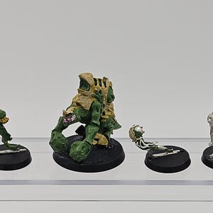 May include: A collection of four miniature gaming figures on black circular bases. The central figure is a green and tan creature with large claws. To its left is a small green appendage, and to its right are two separate silver-colored limbs.