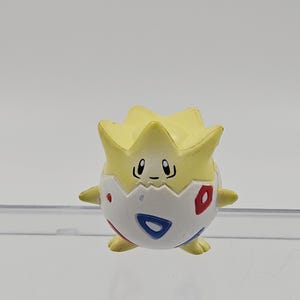 May include: A Togepi figurine, a Pokémon character, is shown. The figure has a white egg-shaped body with red and blue accents. Its head is yellow and spiky, with a small, smiling face. The figurine is on a clear shelf.