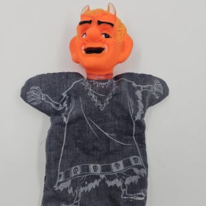 May include: A vintage devil hand puppet with an orange face, yellow hair, and small horns. The puppet's body is a dark gray fabric with a white skeleton design. The puppet has a red collar.