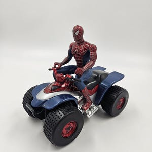 May include: A Spider-Man action figure riding a blue, red, and silver ATV. The ATV has large black tires with red accents. The Spider-Man figure is in a classic red and blue suit.
