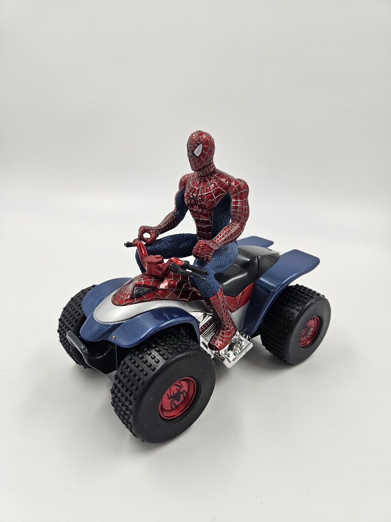 May include: A Spider-Man action figure riding a blue, red, and silver ATV. The ATV has large black tires with red accents. The Spider-Man figure is in a classic red and blue suit.