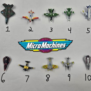 Puede incluir: Una colección de diez aviones de juguete Micro Machines en varios colores y diseños. Los aviones están numerados del 1 al 10 e incluyen una variedad de aviones de combate y otras aeronaves. El logotipo de Micro Machines también es visible.