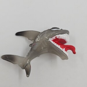 May include: A gray toy shark with a wide open mouth, revealing white teeth and a red tongue. The shark's fins and tail are a darker shade of gray. The toy is set against a plain white background.