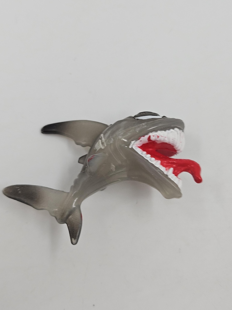 May include: A gray toy shark with a wide open mouth, revealing white teeth and a red tongue. The shark's fins and tail are a darker shade of gray. The toy is set against a plain white background.