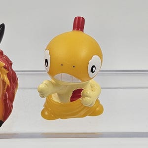 May include: A small yellow and red plastic figurine of a cartoon character. The character has a large head, small arms, and a red crest on top of its head.