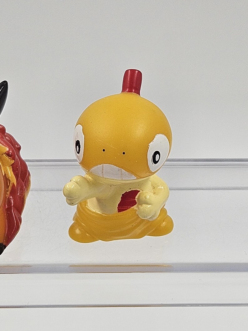 May include: A small yellow and red plastic figurine of a cartoon character. The character has a large head, small arms, and a red crest on top of its head.