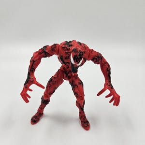May include: A red action figure of a character with a white mouth and black markings. The figure has an open stance with outstretched arms and fingers. The figure is standing on a white surface.
