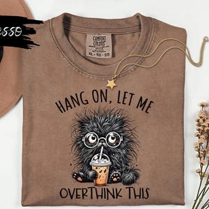 Puede incluir: Camiseta color espresso con el texto "Hang On, Let Me Overthink This" y un monstruo de dibujos animados con gafas sosteniendo una bebida. También es visible un collar de oro con un colgante de estrella.