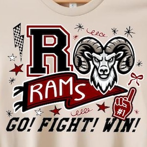 Könnte beinhalten: Beiges T-Shirt mit einem Grafikdesign mit einem Widderkopf, dem Wort "RAMS" und dem Satz "GO! FIGHT! WIN!". Das Design umfasst Sterne, einen Blitz und einen Schaumstoff-Finger. Das Shirt hängt an einem Holzbügel.