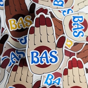 May include: A collection of stickers featuring a hand design with the word "BAS" in blue letters. The hand has a tan base color with a maroon circle on the palm and maroon fingernails. The stickers are scattered on a white surface.