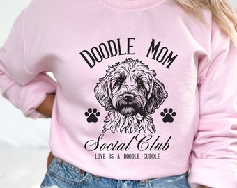 Puede incluir: Sudadera rosa con una ilustración de un perro caniche negro y el texto "Doodle Mom Social Club Love is a doodle cuddle."
