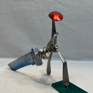 May include: A whimsical sculpture made from recycled silverware, featuring a figure with a red spoon hat standing on a green base. The figure is holding a silver knife.