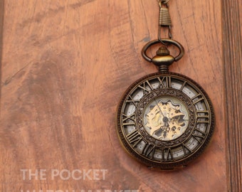 May include: A gold-toned pocket watch with Roman numeral markings on the face and an intricate, exposed gear mechanism. The watch is hanging from a chain on a wooden background.