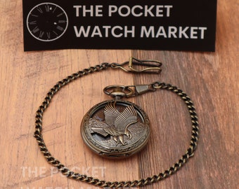 May include: A gold-toned pocket watch with an eagle design on the face. The watch is attached to a gold-toned chain.