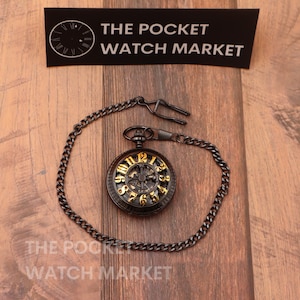Op de afbeelding: Een zwart zakhorloge met een gouden wijzerplaat en Romeinse cijfers. Het horloge heeft een skeletontwerp, waardoor het binnenwerk zichtbaar is. Het is bevestigd aan een zwarte ketting.