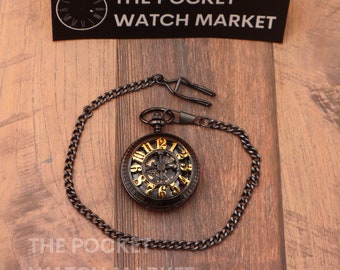 May include: A black pocket watch with a gold face and Roman numeral markings. The watch has a skeletonized design, revealing the inner workings. It is attached to a black chain.