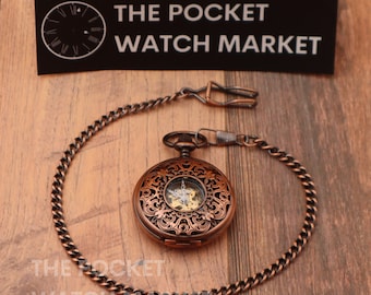 May include: A copper pocket watch with an intricate, open-faced design and a chain. The watch face has Roman numerals and a black background. The watch is on a wooden surface with a black sign that reads "THE POCKET WATCH MARKET".