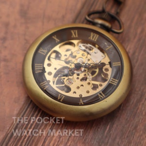 Puede incluir: Un reloj de bolsillo dorado con un diseño esqueletizado, que revela el intrincado funcionamiento interno del reloj. La esfera del reloj tiene marcadores de hora con números romanos y un fondo negro.