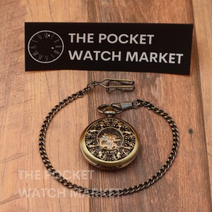 May include: A gold pocket watch with a skeletonized face and intricate engravings. The watch is attached to a gold chain with a clasp.