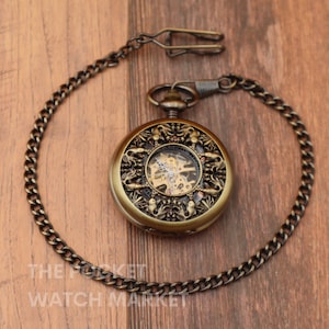 May include: A gold-toned pocket watch with a skeletonized movement and intricate floral design. The watch is attached to a chain.