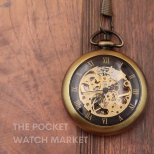 Puede incluir: Un reloj de bolsillo dorado con una cara transparente que revela el intrincado funcionamiento interno del reloj. El reloj tiene marcas de números romanos y un fondo negro.