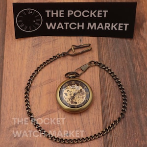 Puede incluir: Un reloj de bolsillo dorado con un movimiento esqueletizado y una cadena. La esfera del reloj tiene marcadores de hora con números romanos y agujas negras. La cadena está unida a un cierre.