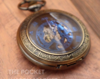 May include: A gold-toned pocket watch with a blue face and an intricate design around the edge. The watch has a clear back that reveals the inner workings of the watch.
