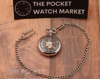 May include: A silver pocket watch with a black face and Roman numeral hour markers. The watch is attached to a silver chain with a clip.