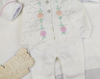 May include: A white knitted baby outfit, including a jacket and pants. The jacket has a Peter Pan collar and is adorned with pink and peach floral embroidery. The outfit is displayed on a white blanket. The wooden sign says "BABY Mel & Kids BOUTIQUE".
