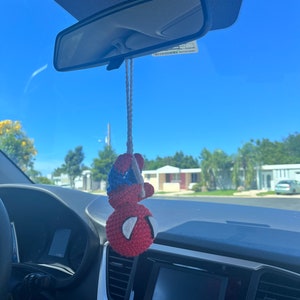 May include: A red and blue crocheted Spiderman hanging from the rearview mirror of a car. The car's interior is visible, including the dashboard and steering wheel.