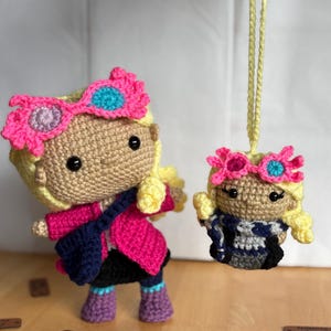 May include: Two crochet dolls, one standing and one hanging. The standing doll has blonde hair, a pink jacket, and a blue purse. The hanging doll has a similar design, with a pink headband and a blue and white outfit. Both dolls have pink and blue accents.
