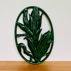 May include: A dark green, oval-shaped wall hanging featuring a detailed relief of corn stalks and ears. The design includes leaves and corn cobs, all in a glossy finish. The decorative piece is mounted on a white wall and a wooden surface.