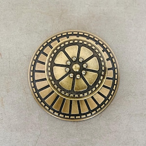 May include: A round brass cabinet knob featuring a tiered design with a central circular element surrounded by eight radiating segments. The outer edge displays a series of rectangular indentations, all finished with a dark, recessed patina.