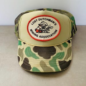 May include: A trucker hat with a camouflage pattern on the bill and a mesh back. The front panel is tan with a red and white oval patch that reads "Lost Dutchman's Mining Association". A gold rope trim separates the front panel from the bill.