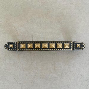 May include: A horizontal drawer pull handle featuring a dark, textured rectangular base with a row of raised, pyramid-shaped brass studs running along the center. The ends of the handle are slightly flared with a decorative, notched edge design.