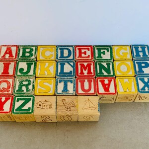 May include: A collection of wooden alphabet blocks arranged in a grid. The blocks feature red, green, yellow, and blue letters from A to Z. Some blocks display illustrations of a house, a rooster, and a sailboat, along with numbers on the sides.