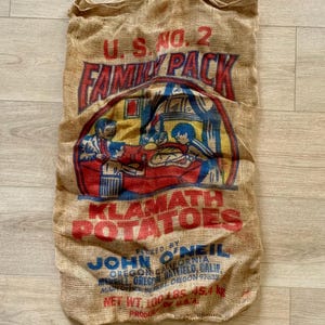 May include: Vintage burlap potato sack with red, blue, and yellow graphics. The sack reads "U.S. No. 2 Family Pack Klamath Potatoes" and "Net Wt. 100 Lbs. 45.4 kg."