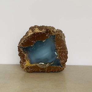 May include: A natural geode with a rough, brown exterior and a smooth, light blue interior. The geode's outer layer has a textured appearance with hints of yellow and brown. The interior has a soft, cloudy appearance.
