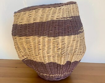 May include: Woven basket with a rounded shape. The basket features alternating bands of light tan and dark purple-brown. The rim is a darker brown. The basket is made of woven material and sits on a wooden surface.