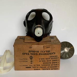 May include: A black gas mask with a white filter is displayed on top of a cardboard box. A white plastic frame and a green filter are also visible. The box has text in Hebrew.