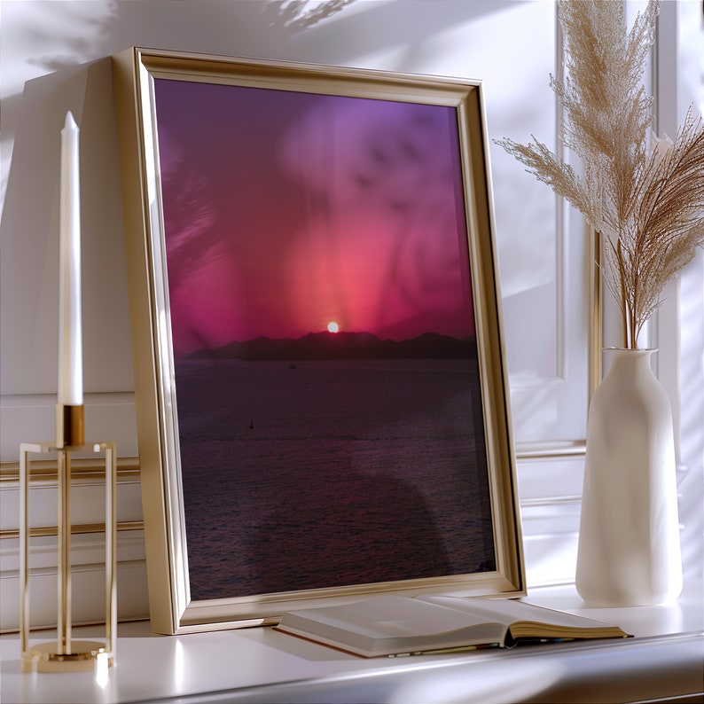 Colorful pink sunset on ocean photography in a golden frame, placed on a modern drawer, along with a long candle and dry leaves in a vase