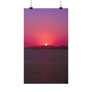 A radiant sun is setting on dark hills and the ocean horizon. The sun has stunning colors and gives the sky beautiful pink and purple hues. The sea is calm and dark in contract with the colorful sky. A single boat crosses the calm sea
Size: 10" x17"