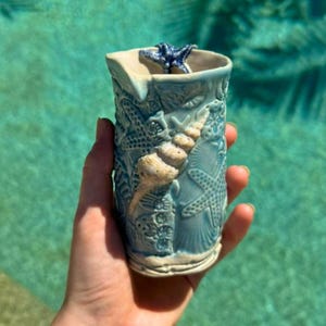 May include: A light blue ceramic tumbler featuring a textured surface with raised relief designs of various seashells and starfish. A dark blue starfish is attached to the top rim, and a large, light-coloured spiral seashell is centred on the front.