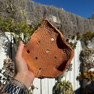 May include: A terracotta-coloured ceramic planter, handmade with a leaf-like shape. The planter has an embossed leaf pattern and several small, circular drainage holes. Small, reflective accents are embedded in the clay.