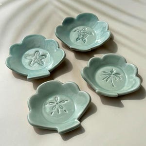 May include: Four pale teal ceramic dishes with a scalloped edge. Each dish features a different embossed design: a starfish, a turtle, a palm tree, and a sand dollar. The dishes are arranged on a light-coloured surface.