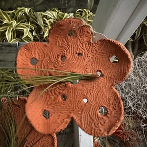 May include: Terracotta flower-shaped ornament with a textured surface and decorative holes. The ornament is a warm orange colour and is suspended by a natural twine. The design features an embossed pattern of swirls and lines.