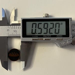 Puede incluir: Un bot&oacute;n marr&oacute;n oscuro con un dise&ntilde;o de pan se mide con un calibrador digital. El bot&oacute;n mide 1,50 cm de di&aacute;metro. El calibrador es plateado y negro, mostrando la medida digitalmente.
