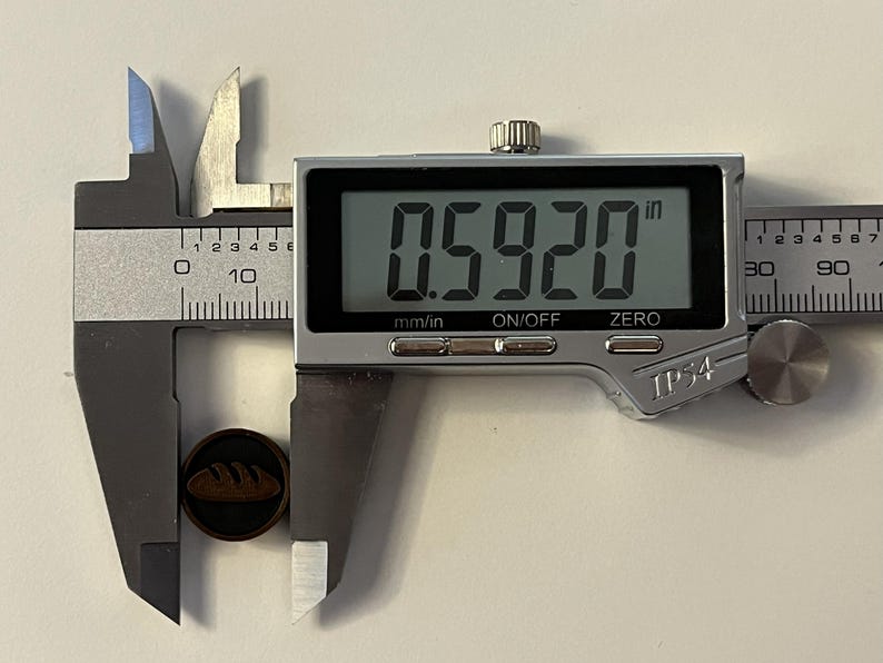 Puede incluir: Un bot&oacute;n marr&oacute;n oscuro con un dise&ntilde;o de pan se mide con un calibrador digital. El bot&oacute;n mide 1,50 cm de di&aacute;metro. El calibrador es plateado y negro, mostrando la medida digitalmente.