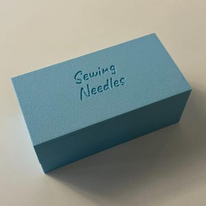 May include: A light blue rectangular box with the words "Sewing Needles" embossed on the top. The box has a textured surface and is sitting on a white surface. The box is a solid color.